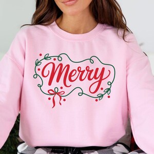 May include: A light pink sweatshirt with the word "Merry" in red script, framed by a green wreath with red berries and a red bow. The sweatshirt is a crew neck style.