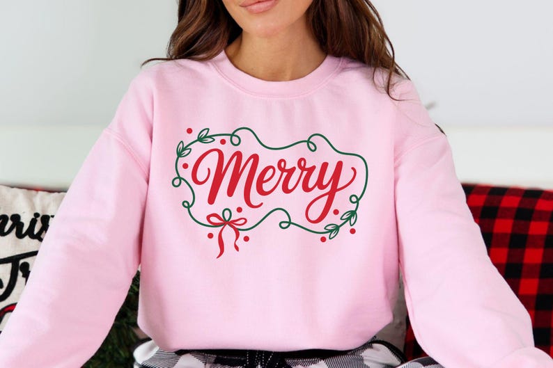May include: A light pink sweatshirt with the word "Merry" in red script, framed by a green wreath with red berries and a red bow. The sweatshirt is a crew neck style.