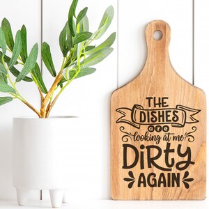 May include: A wooden cutting board with a handle, featuring the text "THE DISHES are looking at me DIRTY AGAIN" in black. A white vase with a green plant is on the left. The image also includes SVG, DXG, PNG, and EPS text.