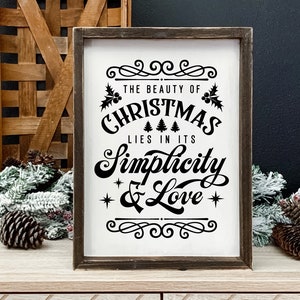 May include: A white sign with a black wood frame and the text "The beauty of Christmas lies in its simplicity & love" with a decorative border and holly sprigs.