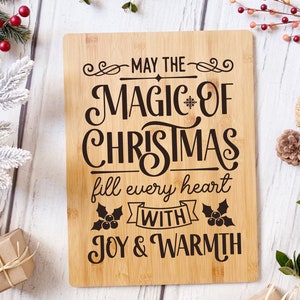 Puede incluir: Una tabla de cortar de madera con el texto "May the magic of Christmas fill every heart with joy & warmth" en letras negras. La tabla est&aacute; rodeada de vegetaci&oacute;n festiva y pi&ntilde;as.