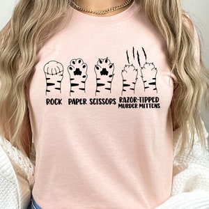 May include: A pink t-shirt with a graphic of a cat paw print in the shape of a rock, paper, scissors hand gesture. The text reads "Rock Paper Scissors Razor-Tipped Murder Mittens".