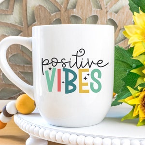 May include: White ceramic mug with a black and colorful design that says "positive vibes". The mug is sitting on a white pedestal with a beaded garland in the foreground.