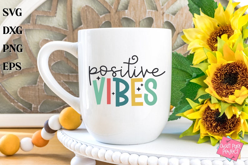 May include: White ceramic mug with a black and colorful design that says "positive vibes". The mug is sitting on a white pedestal with a beaded garland in the foreground.