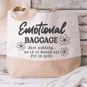 May include: A white canvas tote bag with brown handles. The bag has black text that reads "Emotional Baggage just kidding... as if it would all fit in here." with a daisy design on either side of the text.