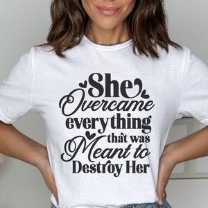 May include: A white t-shirt with black text that reads "She Overcame everything that was Meant to Destroy Her".