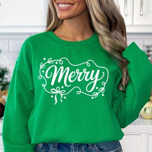May include: A green sweatshirt with the word "Merry" in white script, framed by a decorative border of leaves and bows. The words "DIGITAL DESIGN" are at the top left. The sweatshirt is a festive holiday item.