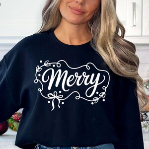May include: A navy blue sweatshirt with the word "Merry" in white script, framed by a decorative floral design. The sweatshirt is a digital design, perfect for holiday apparel.