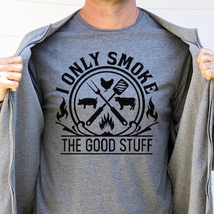Może przedstawiać: Szary T-shirt z czarnym nadrukiem graficznym, na kt&oacute;rym widnieje napis "I only smoke the good stuff" w okręgu, w kt&oacute;rym znajdują się: kurczak, świnia, krowa i skrzyżowane narzędzia do grillowania.