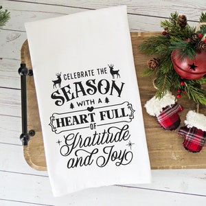 May include: White kitchen towel with black text that reads "Celebrate the Season with a Heart Full of Gratitude and Joy". The text is surrounded by a decorative border with a heart and small trees. Two deer silhouettes are above the text.