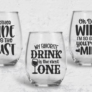 May include: Three wine glasses with funny sayings printed on them. The first glass says "Another Wine Bites the Dust". The second glass says "My Favorite Drink is the Next One". The third glass says "Oh Dear Wine I'm So Glad You're All Mine".
