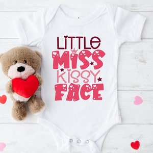 May include: White baby onesie with pink and red text that reads "Little Miss Kissy Face". The text is decorated with hearts and stars. The onesie is on a white wooden background with a brown teddy bear holding a red heart.