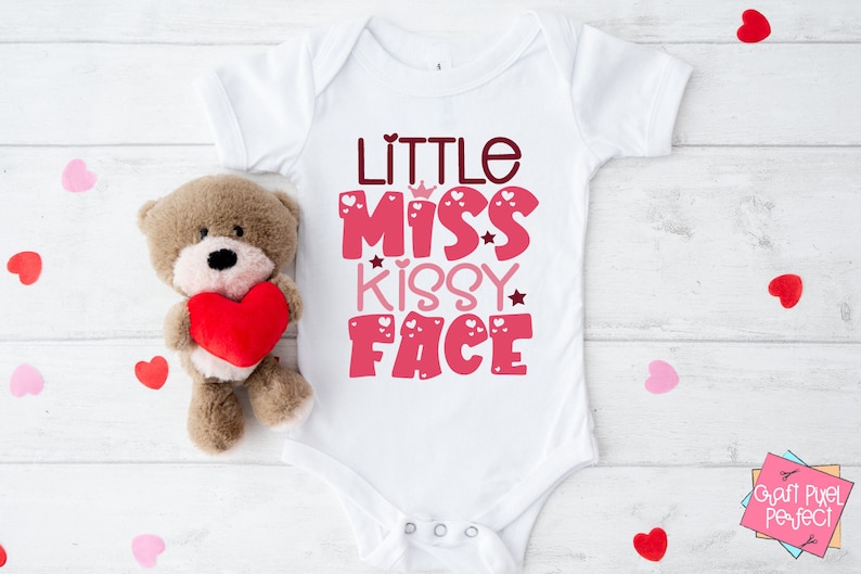 May include: White baby onesie with pink and red text that reads "Little Miss Kissy Face". The text is decorated with hearts and stars. The onesie is on a white wooden background with a brown teddy bear holding a red heart.