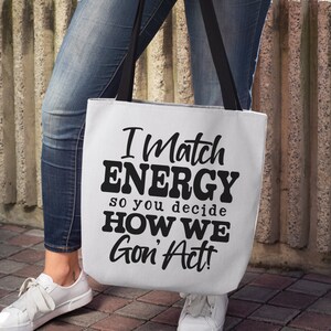 May include: White tote bag with black handles featuring the text "I Match ENERGY so you decide HOW WE Gon' Act!". The bag is held by a person wearing blue jeans and white sneakers. Digital files are also available.