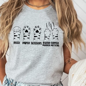 May include: A gray t-shirt with a graphic of cat paws in the shape of rock, paper, scissors, and razor-tipped murder mittens. The text "Rock Paper Scissors Razor-Tipped Murder Mittens" is printed below the graphic.