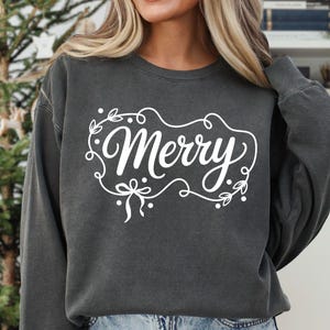 May include: A dark gray sweatshirt with the word "Merry" in white script, framed by a decorative border of leaves and bows. The design is centered on the chest. The sweatshirt is part of a digital design collection.