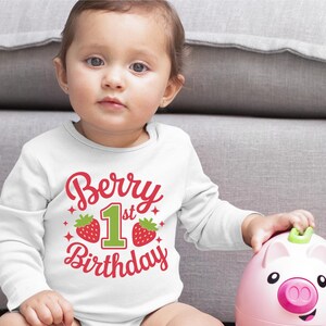 May include: A white long-sleeved shirt with the text "Berry 1st Birthday" in red and green. The number 1 is green and surrounded by two red strawberries.