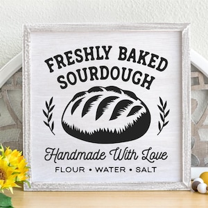 May include: A framed wooden sign with the text "FRESHLY BAKED SOURDOUGH" above a graphic of a loaf of bread. Below the bread graphic, the sign reads "Handmade With Love" and lists "FLOUR &bull; WATER &bull; SALT". The sign is white with black text.