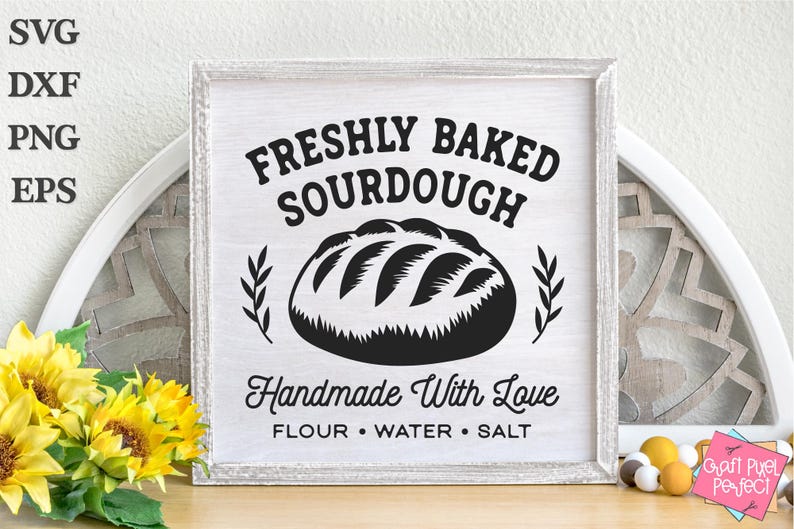 May include: A framed wooden sign with the text "FRESHLY BAKED SOURDOUGH" above a graphic of a loaf of bread. Below the bread graphic, the sign reads "Handmade With Love" and lists "FLOUR &bull; WATER &bull; SALT". The sign is white with black text.