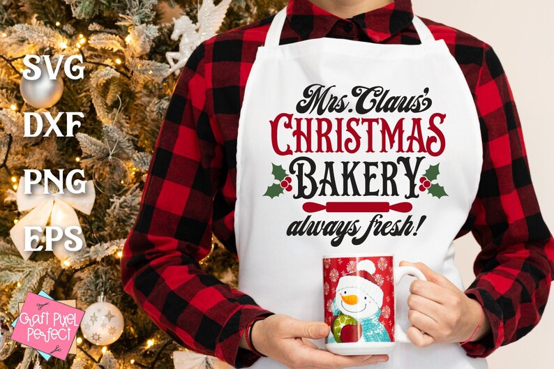 K&ouml;nnte beinhalten: Eine wei&szlig;e Sch&uuml;rze mit dem Text "Mrs. Claus's Christmas Bakery always fresh!" in schwarzer und roter Schrift. Die Sch&uuml;rze hat eine rote Nudelholzgrafik und gr&uuml;nen Stechpalmen mit roten Beeren. Eine Person tr&auml;gt die Sch&uuml;rze und h&auml;lt eine wei&szlig;e Tasse mit einem Schneemann-Design.