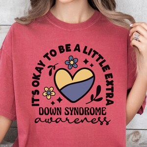 May include: A red t-shirt with a black circle design that says "It's okay to be a little extra Down Syndrome awareness" with a yellow and blue heart and floral accents.