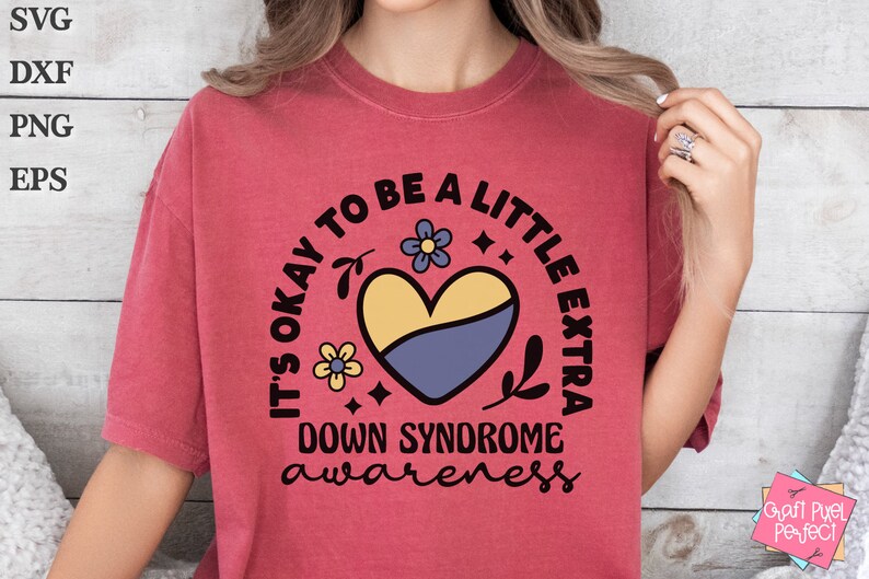 May include: A red t-shirt with a black circle design that says "It's okay to be a little extra Down Syndrome awareness" with a yellow and blue heart and floral accents.