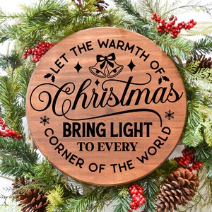 May include: A round wooden sign with the text "Let the warmth of Christmas bring light to every corner of the world". The sign is decorated with holly, snowflakes, and bells. The sign is surrounded by a green Christmas wreath with red berries and pine cones.