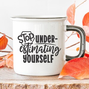 May include: White ceramic mug with a black handle. The mug features a black graphic that says "Stop Under-estimating Yourself". The mug is resting on a wooden surface with autumn leaves surrounding it.