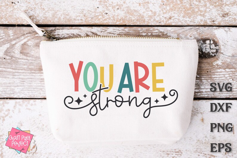 May include: A white canvas zippered pouch with the text "YOU ARE strong" in colorful letters and a black swirl design with stars. The text is in a playful font. The pouch is on a light wood background.