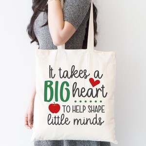 May include: A white tote bag with the text "It takes a BIG heart ... TO HELP SHAPE little minds" in green and black lettering. A red heart and a red apple are also included in the design.