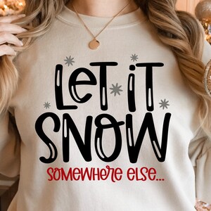 May include: A white sweatshirt with the text "Let it snow... somewhere else." in black lettering with gray snowflakes. The text "somewhere else" is in red.
