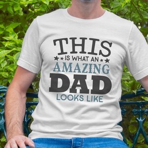 May include: A white t-shirt with the text "THIS IS WHAT AN AMAZING DAD LOOKS LIKE" printed in black and blue lettering.