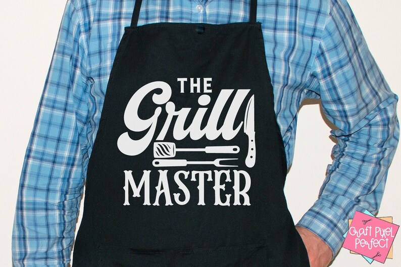 May include: Black apron with the text "THE Grill MASTER" in white. The design includes illustrations of a spatula, fork, and knife. The apron is worn over a blue plaid shirt.