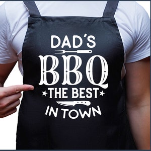 May include: A black apron with white text that reads "Dad's BBQ *The Best* In Town". The text is stylized with a vintage font and includes a grill fork and knife graphic.