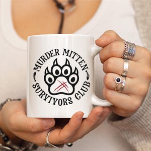 Pu&ograve; includere: Tazza in ceramica bianca con un design grafico nero e rosso. Il design presenta un'impronta di zampa con il testo "Murder Mitten Survivors Club".