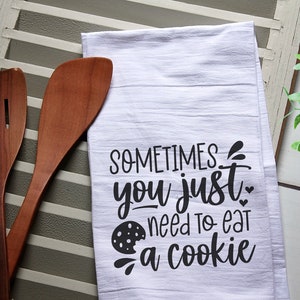 May include: White kitchen towel with black text that reads "Sometimes you just need to eat a cookie" with a black and white cookie graphic. The towel is on a white wooden surface with two wooden spoons.