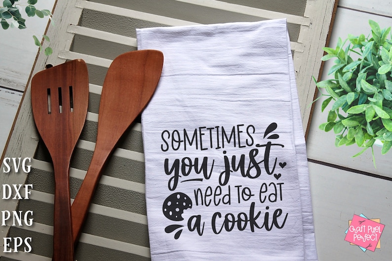May include: White kitchen towel with black text that reads "Sometimes you just need to eat a cookie" with a black and white cookie graphic. The towel is on a white wooden surface with two wooden spoons.