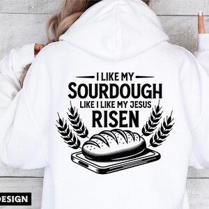 May include: A white hoodie with black text that reads "I like my sourdough like I like my Jesus risen". The text is surrounded by wheat stalks and a black and white illustration of a loaf of sourdough bread on a cutting board.