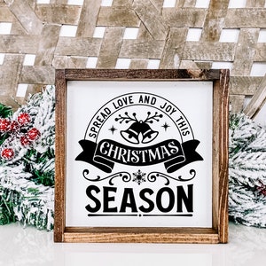 May include: A white wooden sign with black text that reads "Spread love and joy this Christmas season". The sign is decorated with a snowflake, bells, and a banner. The sign is framed in a brown wooden frame.