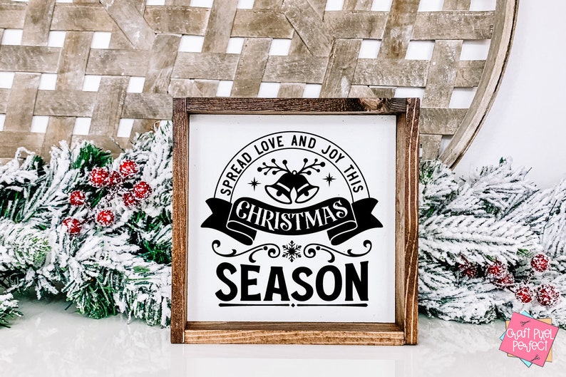 May include: A white wooden sign with black text that reads "Spread love and joy this Christmas season". The sign is decorated with a snowflake, bells, and a banner. The sign is framed in a brown wooden frame.