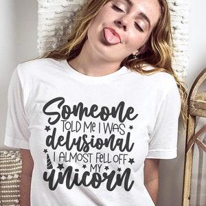 May include: A white t-shirt with black text that reads "Someone told me I was delusional I almost fell off my unicorn". The text is surrounded by small stars. The shirt is being worn by a person with long blonde hair.