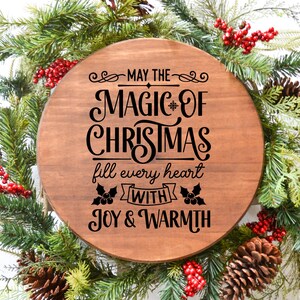 Puede incluir: Un letrero redondo de madera con el texto "May the magic of Christmas fill every heart with joy & warmth" en letras negras. El letrero est&aacute; rodeado de una guirnalda verde con bayas rojas y pi&ntilde;as.