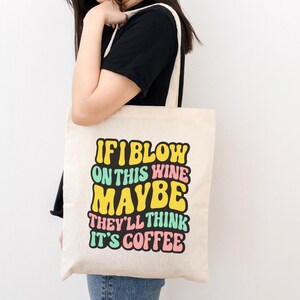 Op de afbeelding: Een witte tote bag met de tekst "If I blow on this wine maybe they'll think it's coffee" in kleurrijk retro-lettertype.