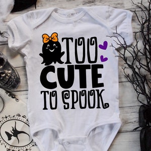 K&ouml;nnte beinhalten: Ein wei&szlig;er Baby-Strampler mit einem schwarzen Geistermotiv und dem Text "Too Cute To Spook" in Schwarz. Der Geist hat eine rosa Schleife und ein fr&ouml;hliches Gesicht. Auf dem Strampler befinden sich zwei lila Herzen.