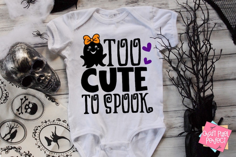 K&ouml;nnte beinhalten: Ein wei&szlig;er Baby-Strampler mit einem schwarzen Geistermotiv und dem Text "Too Cute To Spook" in Schwarz. Der Geist hat eine rosa Schleife und ein fr&ouml;hliches Gesicht. Auf dem Strampler befinden sich zwei lila Herzen.