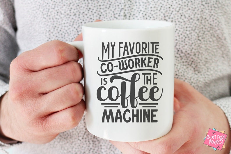 May include: White ceramic coffee mug with the text "MY FAVORITE CO-WORKER IS THE coffee MACHINE" in black lettering. The mug is held by a person. The mug has a handle on the right side. The mug is a standard size.