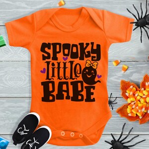 May include: Orange baby onesie with the text "Spooky Little Babe" in black with a purple heart and a ghost with a bow.