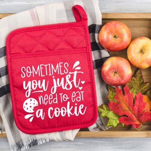 May include: Red quilted hot pad with the text "SOMETIMES you just need to eat a cookie" in white. A cookie is pictured next to the text. The hot pad is on a wooden tray with a checkered cloth, apples, and fall leaves.