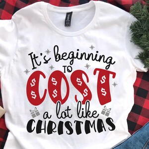 May include: White t-shirt with the text "It's beginning to cost a lot like Christmas" in black and red lettering. The word "cost" is surrounded by dollar signs.