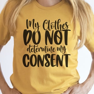 Puede incluir: Una camiseta amarilla con el texto "My Clothes Do Not Determine My Consent" en letras negras.